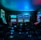 Dimly lit gaming arcade featuring a variety of brightly illuminated slot machines and video gaming screens. Neon signs and colorful displays create a vibrant atmosphere. The arrangement of gaming machines suggests an inviting space for entertainment, with chairs positioned for players.