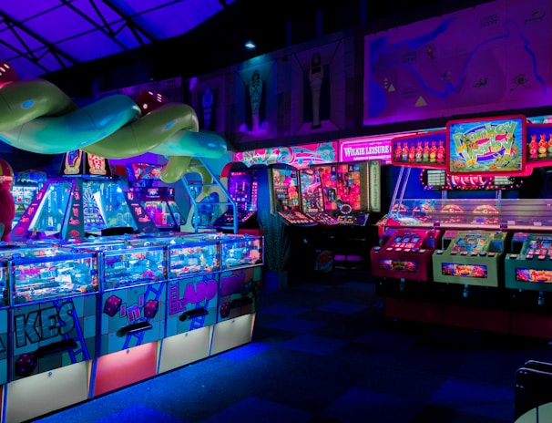An indoor arcade filled with vibrant gaming machines lit with neon blue and pink lights. The scene includes various arcade games with flashing screens and colorful displays, and a large overhead structure resembling a green snake. The background shows illuminated signs and themed decor, contributing to an immersive entertainment atmosphere.
