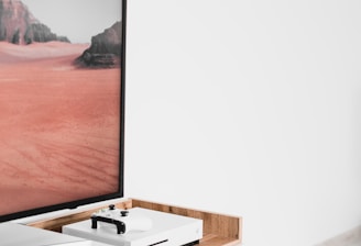 A modern interior setting featuring a white entertainment console with wooden accents. On the console, there is a gaming console with a controller on top. A large framed picture of a desert landscape with red sand and rocky formations is positioned next to the console.