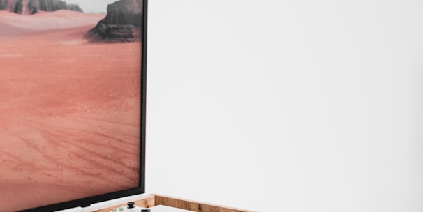 A modern interior setting featuring a white entertainment console with wooden accents. On the console, there is a gaming console with a controller on top. A large framed picture of a desert landscape with red sand and rocky formations is positioned next to the console.