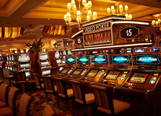 A casino floor featuring rows of brightly lit video poker machines. The area is illuminated by elegant chandeliers, with a display board showing a large sum of money, suggesting a possible jackpot. The rich decor includes luxurious carpets and comfortable seating, adding to the opulent atmosphere.