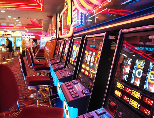 A brightly lit casino interior featuring a row of colorful slot machines. The neon lights create a vibrant atmosphere, with patterned carpeting adding to the decor. Stools are aligned neatly in front of each machine.