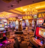A vibrant casino interior filled with rows of colorful slot machines, each illuminated with bright lights and displays. The ceiling is adorned with elegant chandeliers and golden drapes, creating a luxurious atmosphere. The patterned carpet adds to the elaborate design of the space.