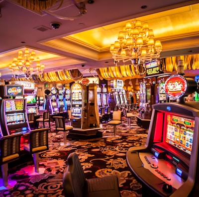 A vibrant casino interior filled with rows of colorful slot machines, each illuminated with bright lights and displays. The ceiling is adorned with elegant chandeliers and golden drapes, creating a luxurious atmosphere. The patterned carpet adds to the elaborate design of the space.