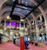 The interior of a large, ornately designed building with arched ceilings and intricate architectural details. The space features gaming machines lined up along the floor, under a high ceiling with a grid pattern. Lighting in purple and white tones enhances the ambiance.