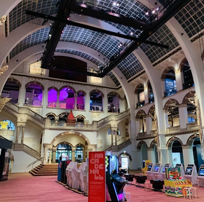 The interior of a large, ornately designed building with arched ceilings and intricate architectural details. The space features gaming machines lined up along the floor, under a high ceiling with a grid pattern. Lighting in purple and white tones enhances the ambiance.