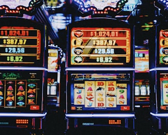 A row of colorful slot machines lit with bright neon lights, showcasing various graphical elements on the screens, indicating different games and jackpots. The atmosphere suggests a lively casino setting, bustling with excitement.