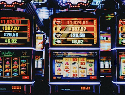 A row of colorful slot machines lit with bright neon lights, showcasing various graphical elements on the screens, indicating different games and jackpots. The atmosphere suggests a lively casino setting, bustling with excitement.