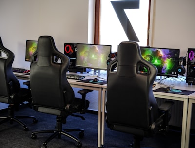 A row of gaming stations equipped with large monitors, gaming chairs, keyboards, and headphones. The setup is organized and ready for gaming, featuring a colorful screen display. The room has a modern and tech-savvy ambiance.