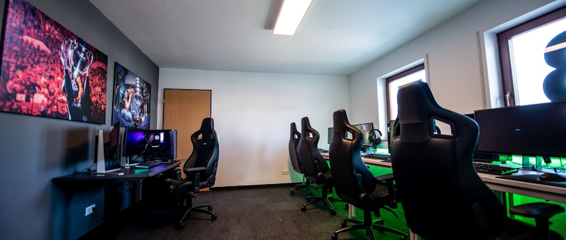 A well-lit gaming room features multiple gaming stations with high-backed chairs and monitors. The room has a minimalist setup with green and black color tones. Large posters adorn the walls, and the room appears clean and organized.
