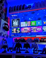 A modern gaming setup featuring a large wall-mounted screen displaying a gaming console interface. The room is decorated with gaming-themed items such as controllers and collectibles. The ambient lighting includes shades of blue and red, creating a vibrant atmosphere.
