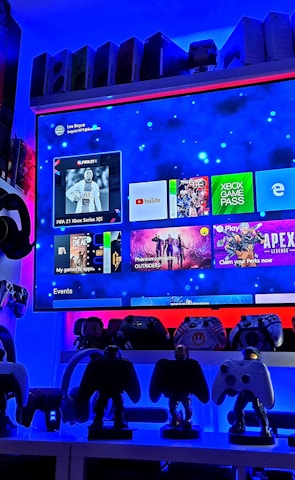 A modern gaming setup featuring a large wall-mounted screen displaying a gaming console interface. The room is decorated with gaming-themed items such as controllers and collectibles. The ambient lighting includes shades of blue and red, creating a vibrant atmosphere.