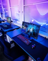 A row of modern gaming setups is arranged neatly along a window-lined wall. Each station includes a high-resolution monitor displaying vivid graphics, a mechanical keyboard with colored lighting, a gaming mouse, and an ergonomic chair. The room is illuminated with ambient blue and purple lights, creating a sleek and futuristic atmosphere.