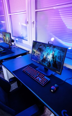 A row of modern gaming setups is arranged neatly along a window-lined wall. Each station includes a high-resolution monitor displaying vivid graphics, a mechanical keyboard with colored lighting, a gaming mouse, and an ergonomic chair. The room is illuminated with ambient blue and purple lights, creating a sleek and futuristic atmosphere.