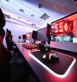 A row of high-end gaming computers on sleek desks, featuring RGB lighting and gaming accessories. The computers are housed in transparent cases with visible components, emitting a red glow. Comfortable gaming chairs with ergonomic design are positioned next to the desks, and the room's ceiling is modern with recessed lighting.