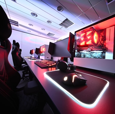 A row of high-end gaming computers on sleek desks, featuring RGB lighting and gaming accessories. The computers are housed in transparent cases with visible components, emitting a red glow. Comfortable gaming chairs with ergonomic design are positioned next to the desks, and the room's ceiling is modern with recessed lighting.
