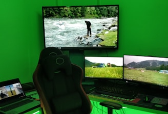 A gaming setup with a large curved monitor showing a scenic image of a river and forest. Several smaller monitors display nature scenes, and a laptop is open showing a different video. A comfortable gaming chair is positioned in front of the setup. The room is illuminated by green lighting, and the desktop tower has colorful RGB fans.