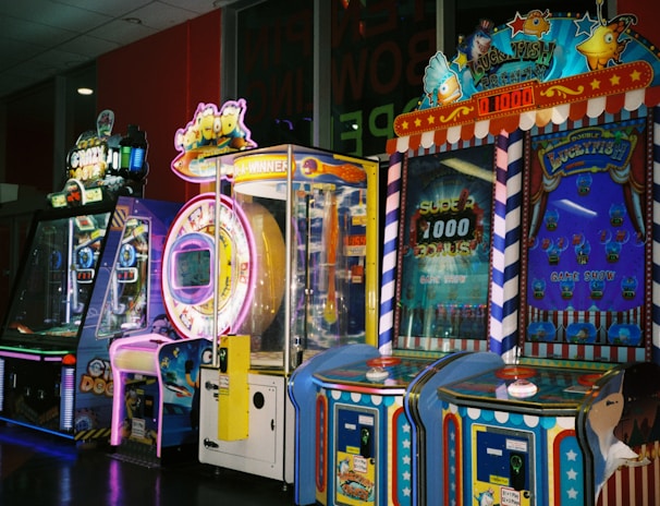 A row of brightly colored arcade game machines is arranged in a line against the wall. Each machine features vibrant lights and designs, with themes ranging from claw games to game show-inspired setups. The area has a playful and inviting atmosphere, suitable for entertainment or amusement centers.