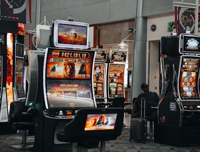 A collection of slot machines is situated inside a building with bright, colorful screens displaying various games and gaming themes. A person is seated nearby, seemingly relaxed with luggage beside them, and the atmosphere is lively with electronic displays.