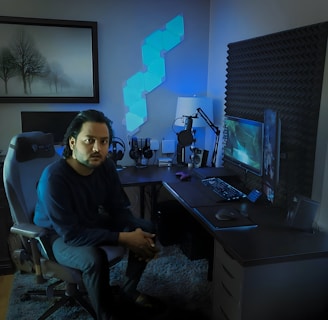 A person is seated on a gaming chair in a dimly lit room with a modern setup that includes a computer, microphone, and multiple screens. The walls are decorated with soundproofing panels and a geometric LED light panel. A framed picture of trees is hung on the wall. The atmosphere suggests a gaming or streaming environment.