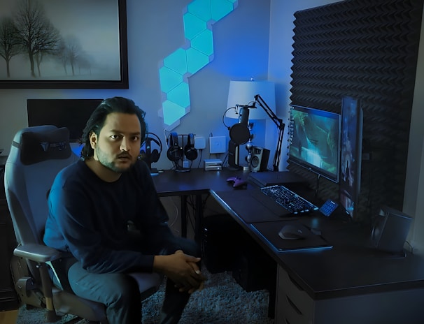 A person is seated on a gaming chair in a dimly lit room with a modern setup that includes a computer, microphone, and multiple screens. The walls are decorated with soundproofing panels and a geometric LED light panel. A framed picture of trees is hung on the wall. The atmosphere suggests a gaming or streaming environment.